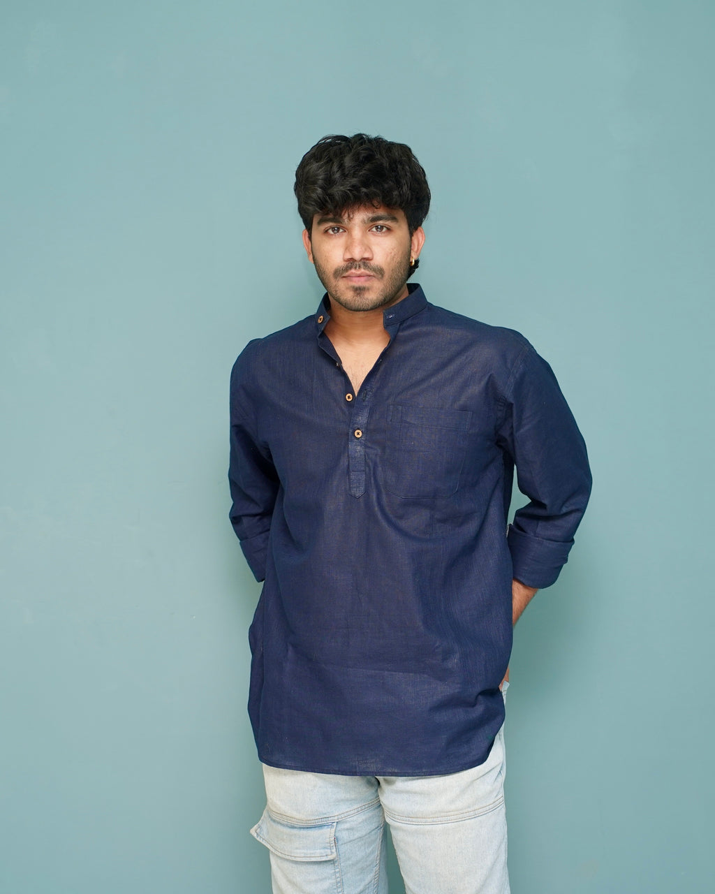 A man wearing a navy blue long-sleeve button-up shirt and light gray jeans, standing against a solid light blue-gray background.