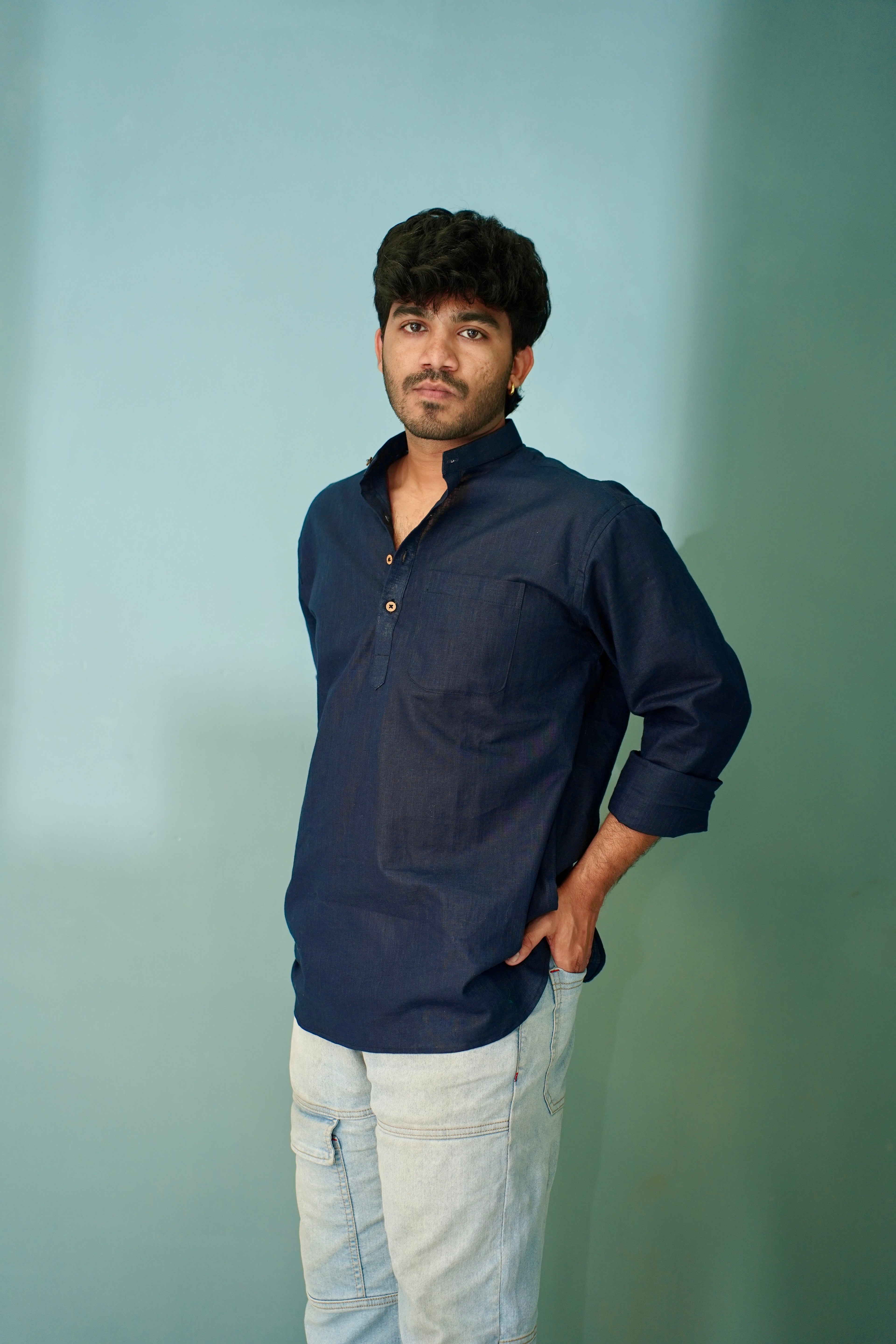 A man wearing a dark navy blue long-sleeve button-up shirt and light beige jeans, standing against a solid light blue-green background.