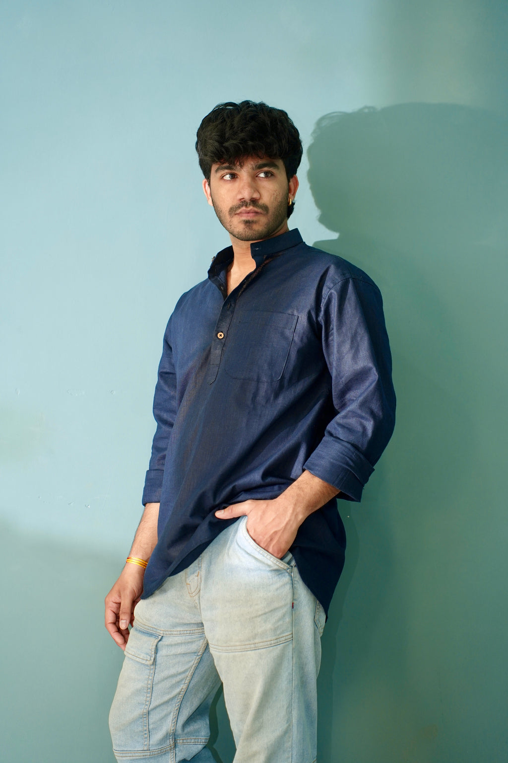 A man wearing a navy blue long-sleeve kurta and light blue jeans, standing against a solid light blue wall with his hand in his pocket.