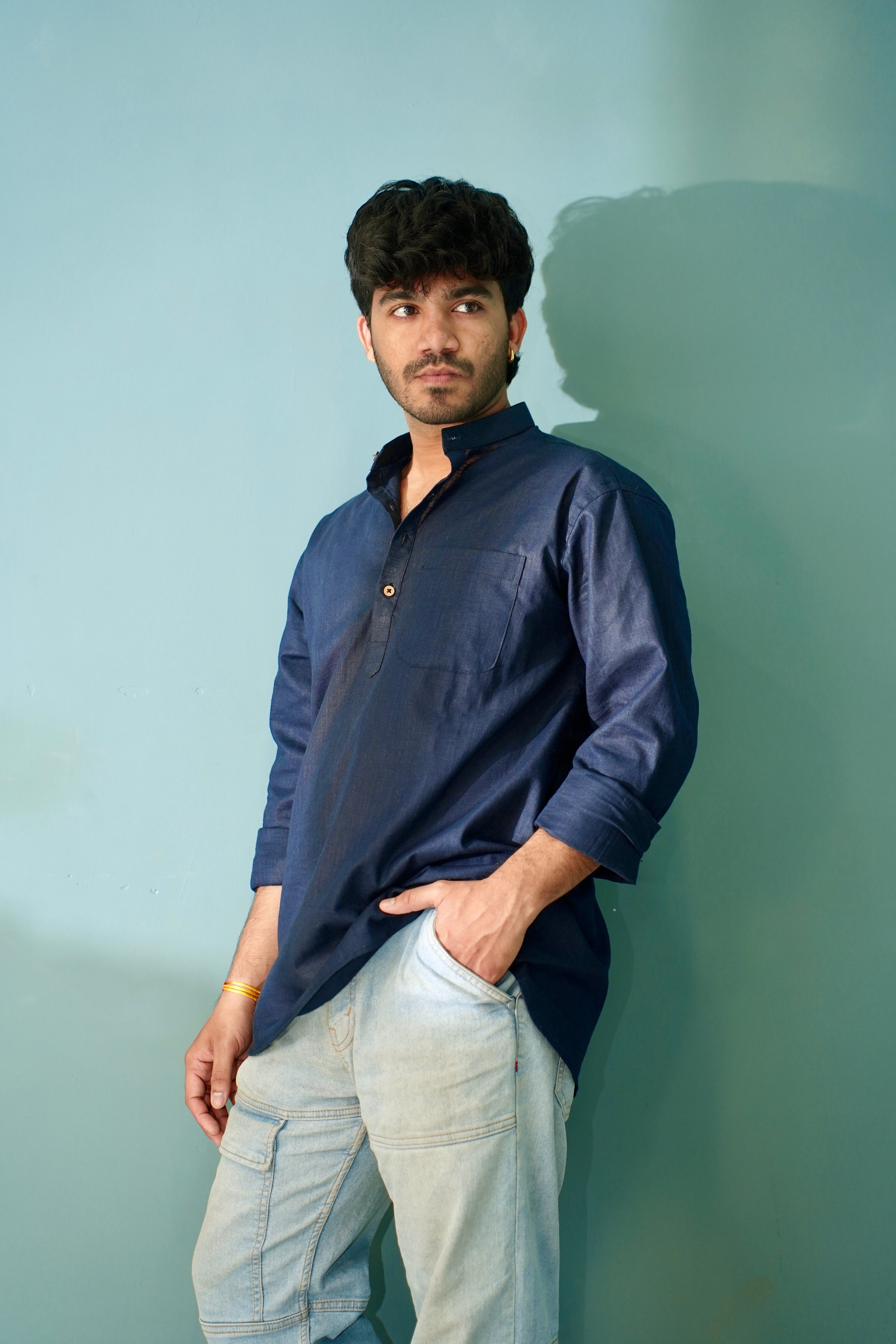 A man wearing a navy blue long-sleeve kurta and light blue jeans, standing against a solid light blue wall with his hand in his pocket.