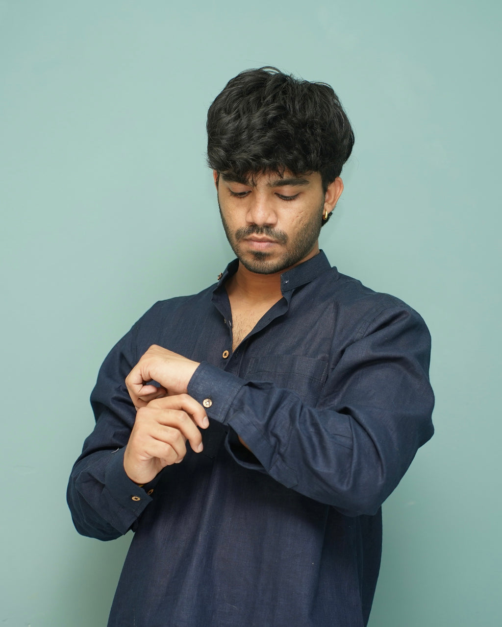 A man wearing a dark navy blue long-sleeve button-up shirt, adjusting the cuff of his right sleeve against a solid light teal background.