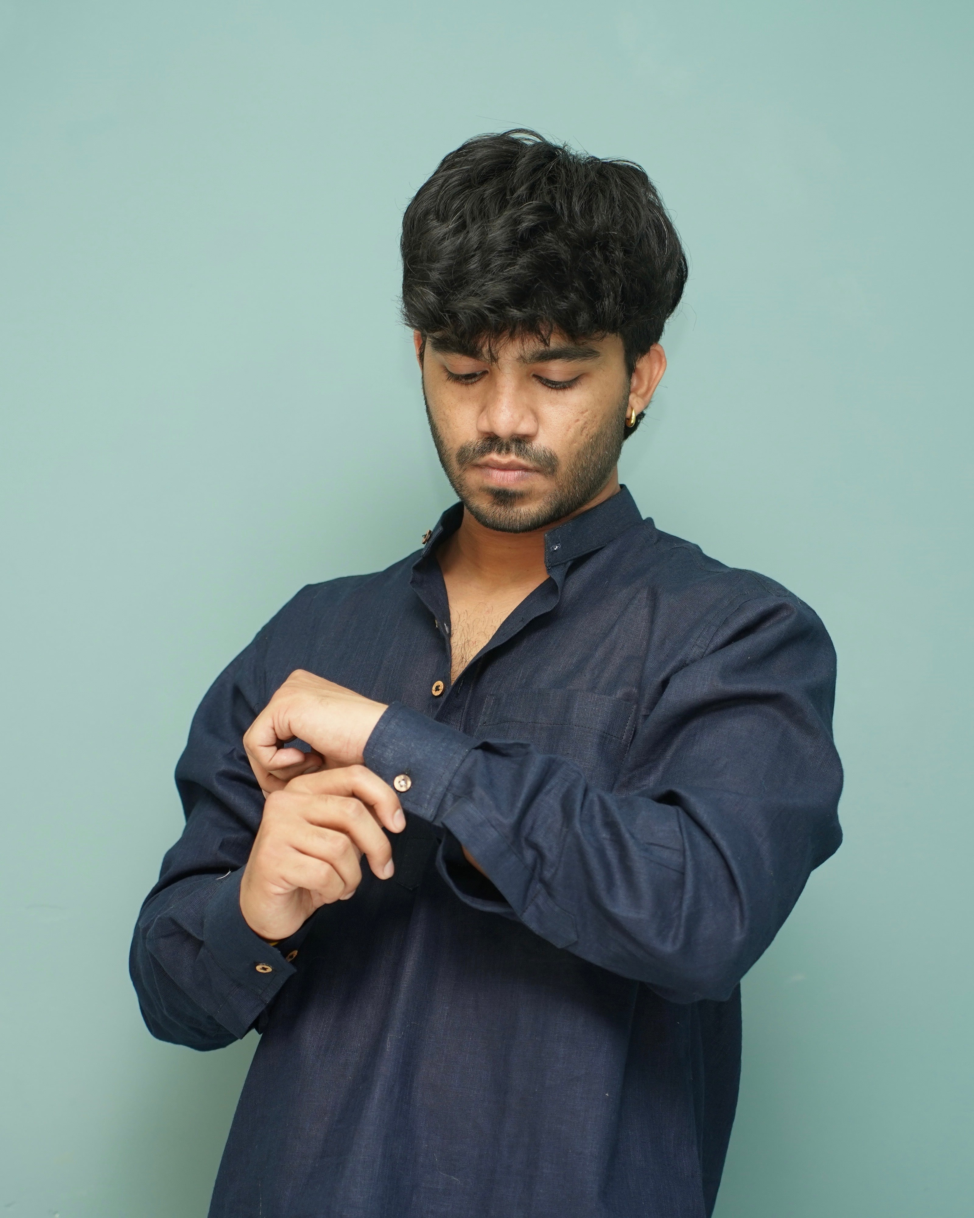 A man wearing a dark navy blue long-sleeve button-up shirt, adjusting the cuff of his right sleeve against a solid light teal background.
