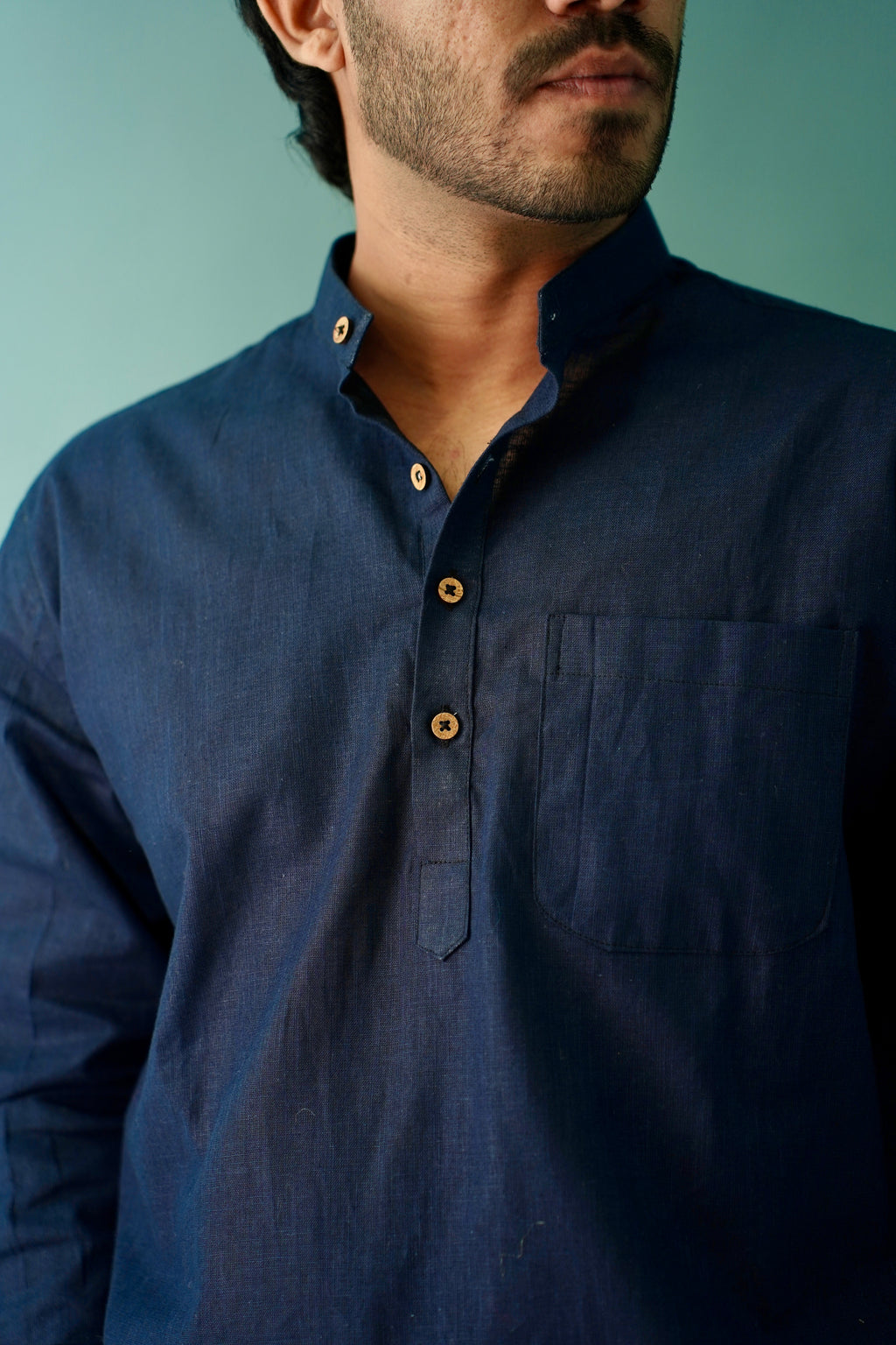 A close-up of a man wearing a dark navy blue long-sleeve linen shirt with a buttoned collar and brown buttons, set against a solid teal background.