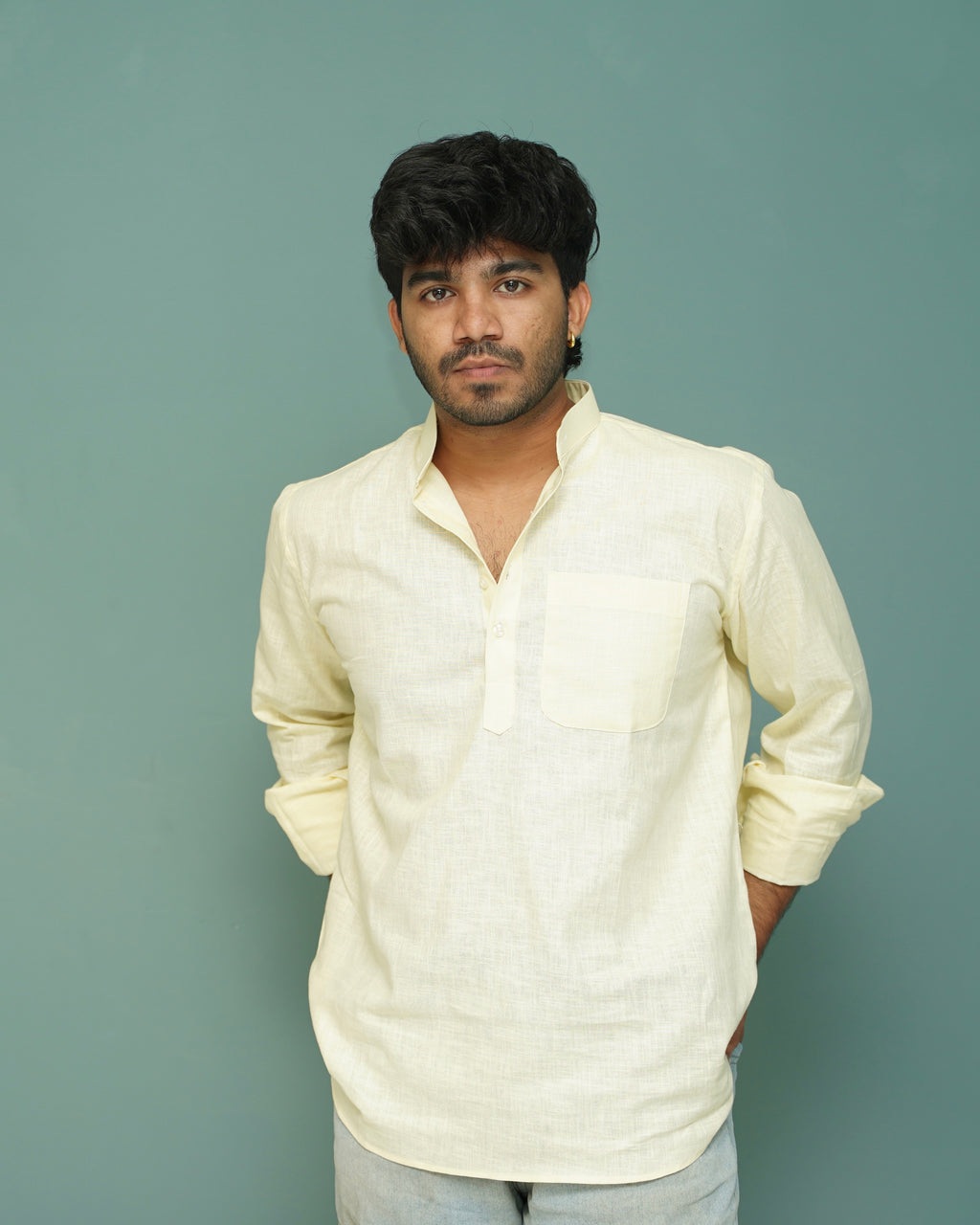 A man wearing a light cream-colored long-sleeve linen shirt with a chest pocket, standing against a solid muted teal background.