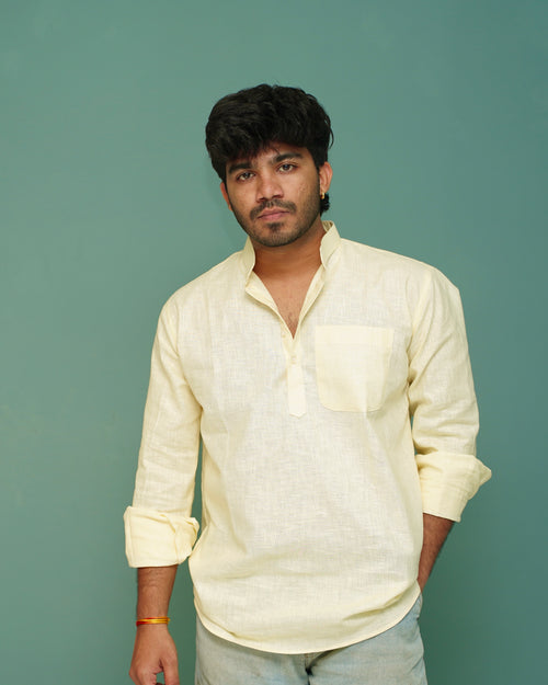 A man wearing a light cream-colored linen shirt with a chest pocket and light gray jeans, standing against a solid teal-green background.