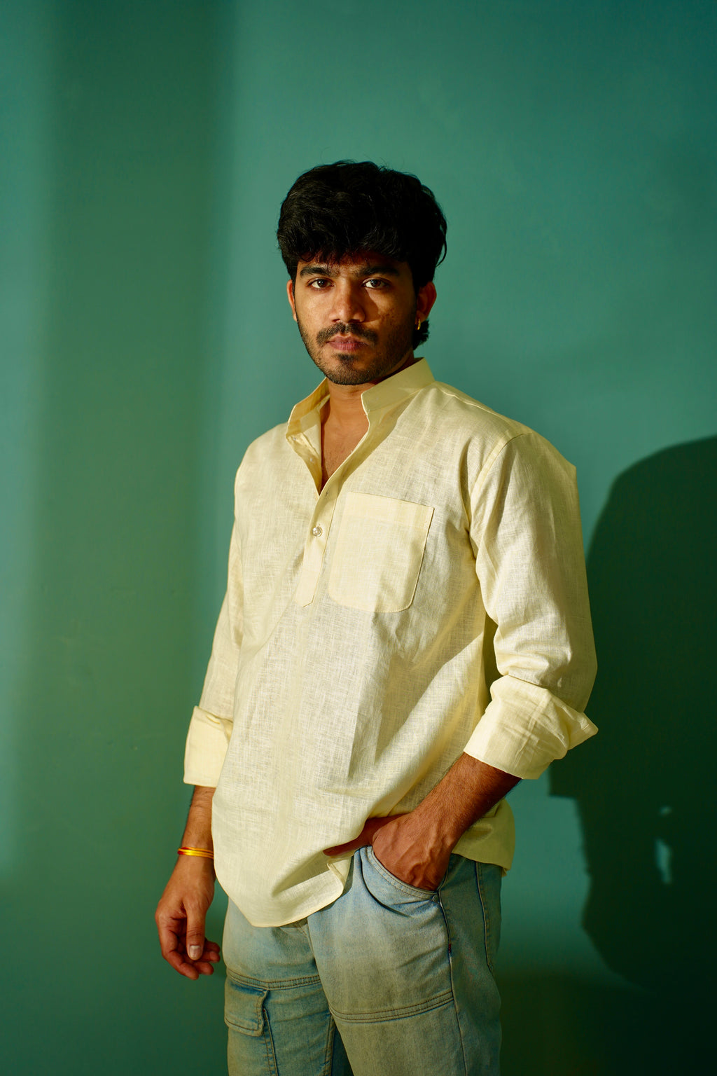 A man wearing a light cream-colored linen shirt and light blue jeans, standing against a solid teal-green background with a soft shadow on the right.