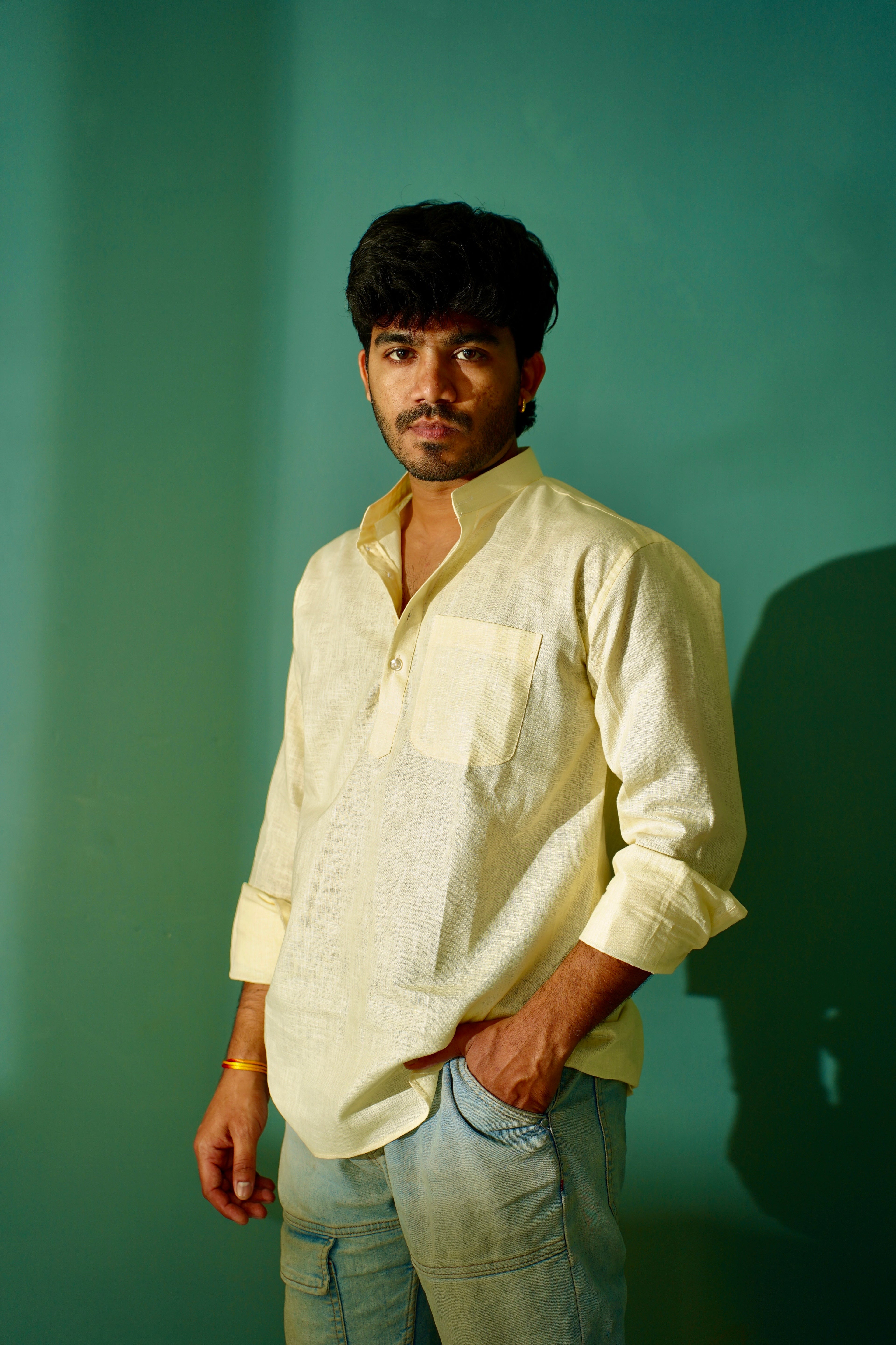 A man wearing a light cream-colored linen shirt and light blue jeans, standing against a solid teal-green background with a soft shadow on the right.