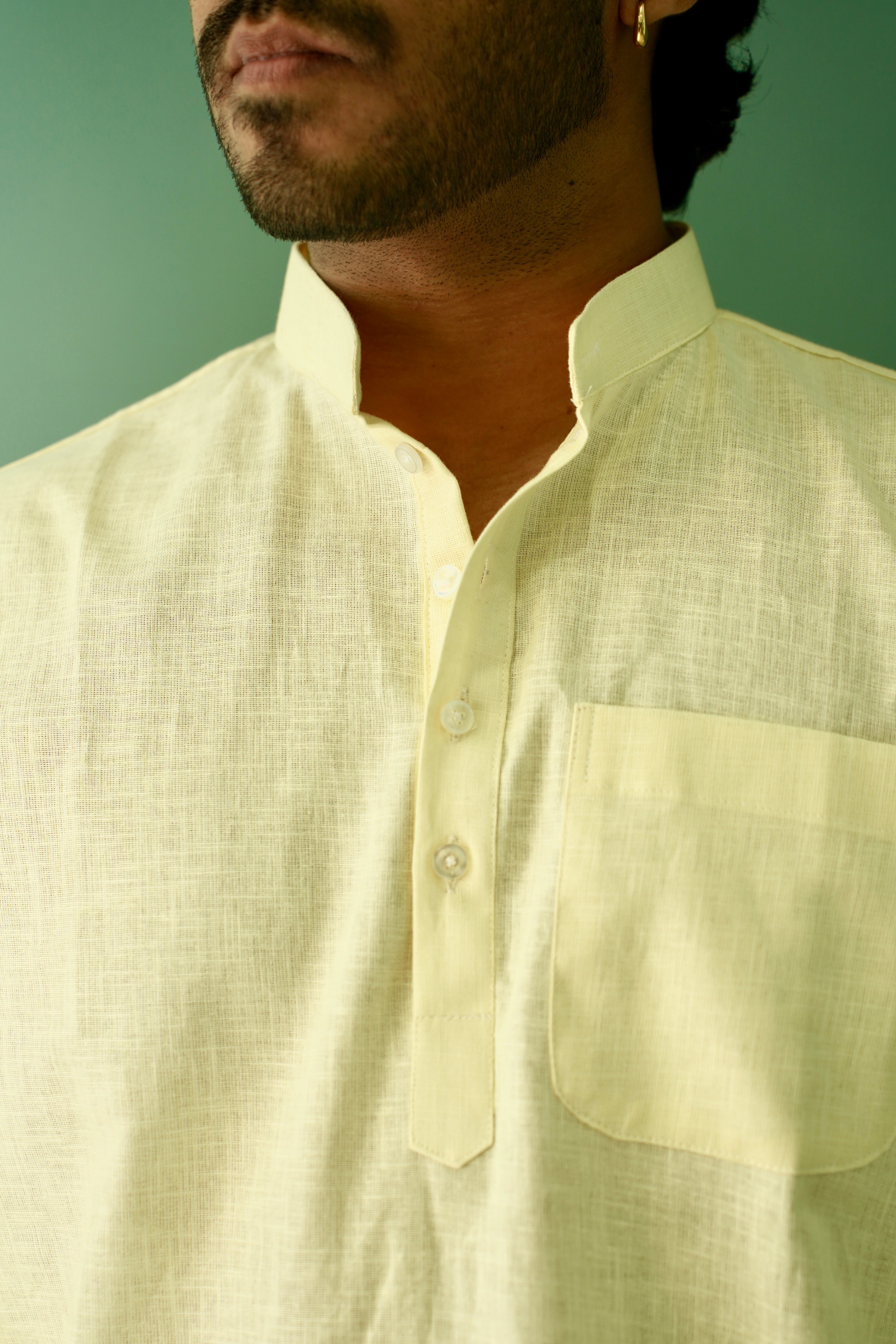 A close-up of a man wearing a pale yellow linen kurta with a mandarin collar, button placket, and a chest pocket, set against a solid green background.