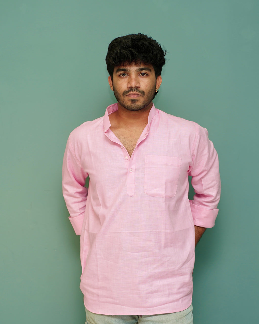 A man wearing a light pink long-sleeve linen shirt with a half-placket and chest pocket, standing against a solid sage green background.