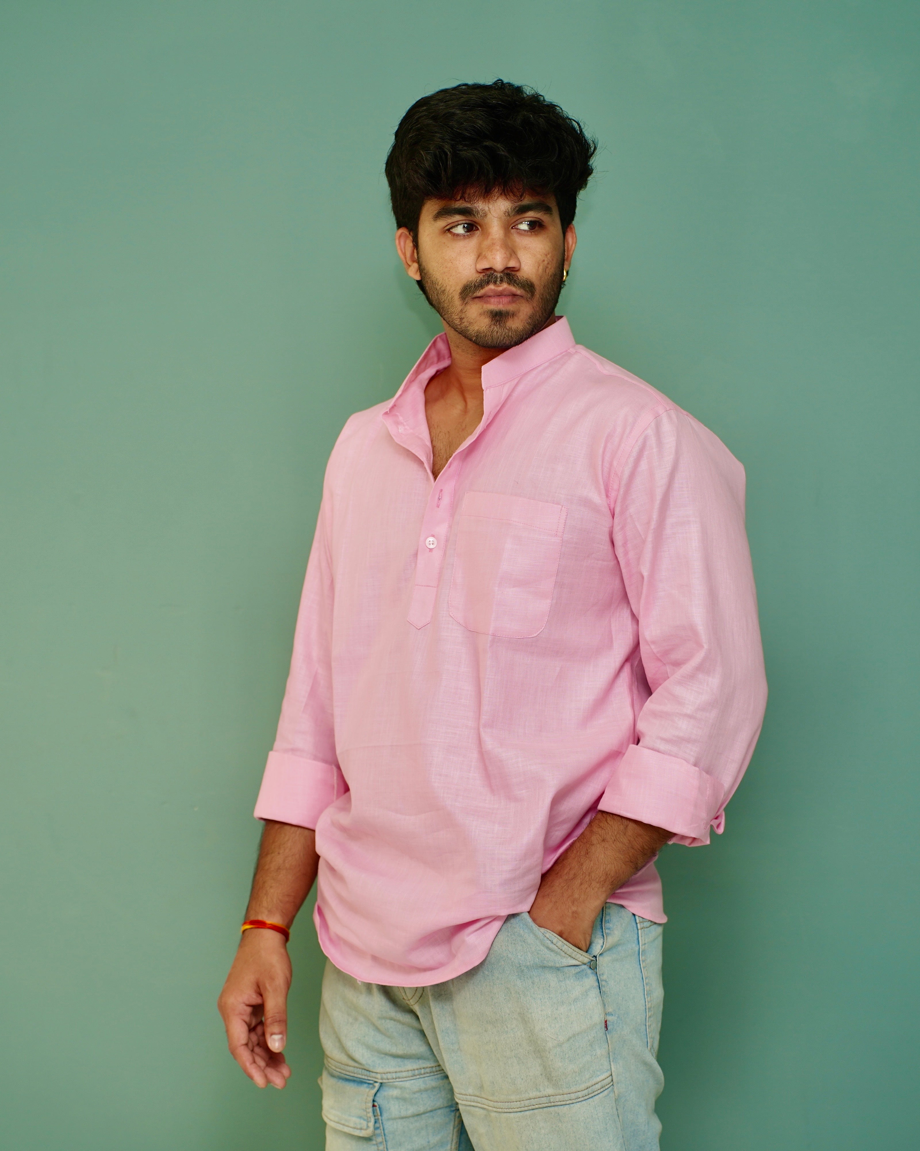A man wearing a light pink long-sleeve linen shirt and light blue jeans, standing against a solid sage green background.