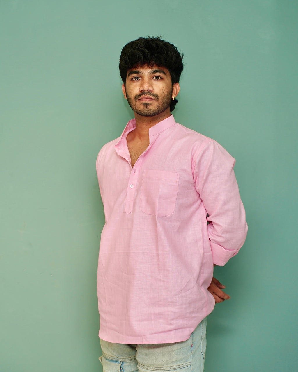 A man wearing a light pink long-sleeve cotton kurta with a chest pocket, paired with light blue jeans, standing against a solid mint green background.