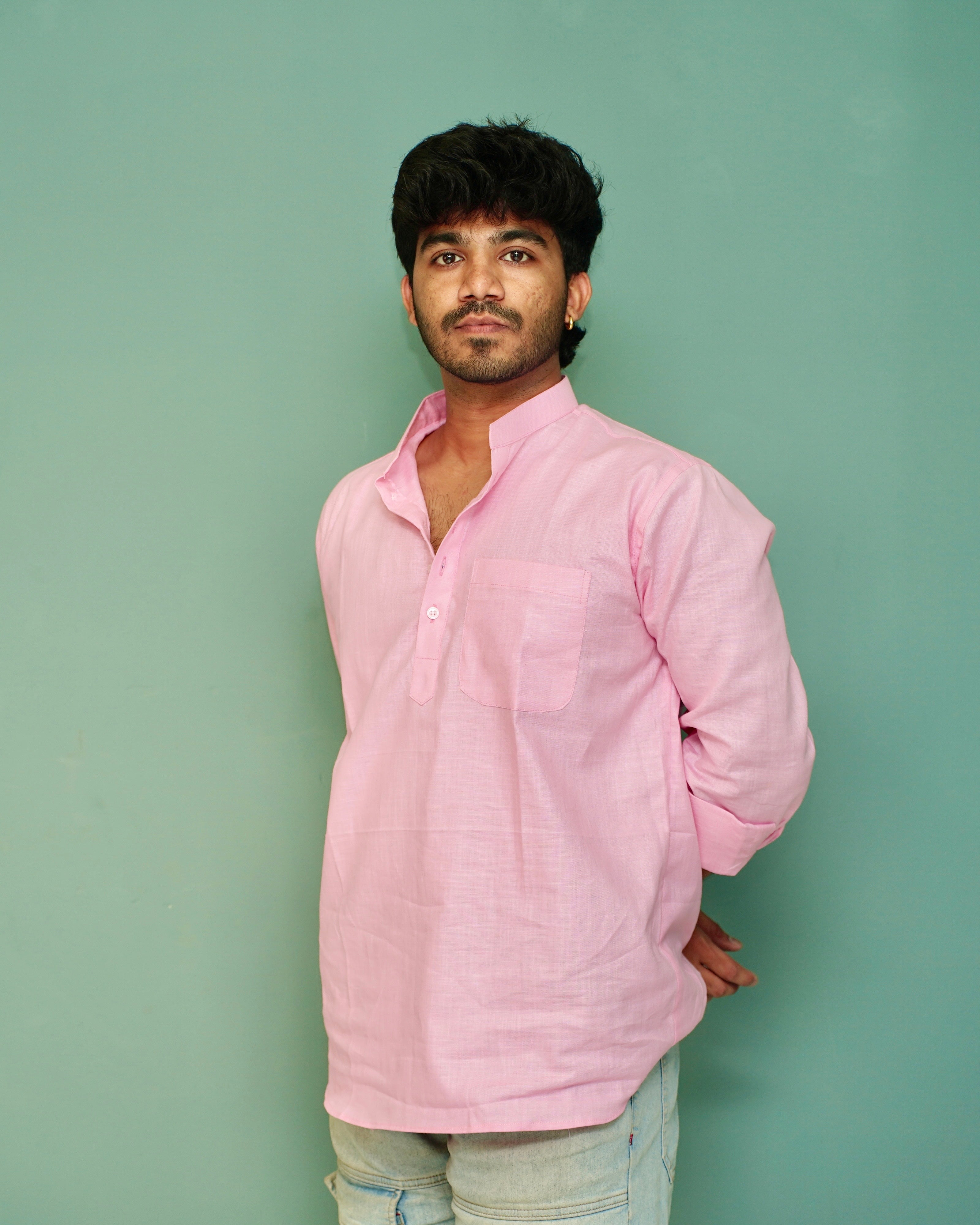A man wearing a light pink long-sleeve cotton kurta with a chest pocket, paired with light blue jeans, standing against a solid mint green background.