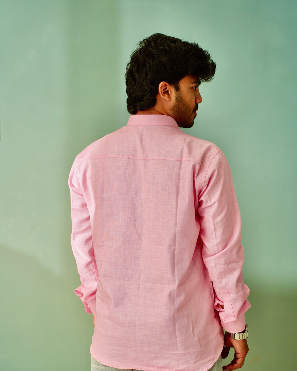A man viewed from behind wearing a light pink long-sleeve linen shirt against a solid mint green background.
