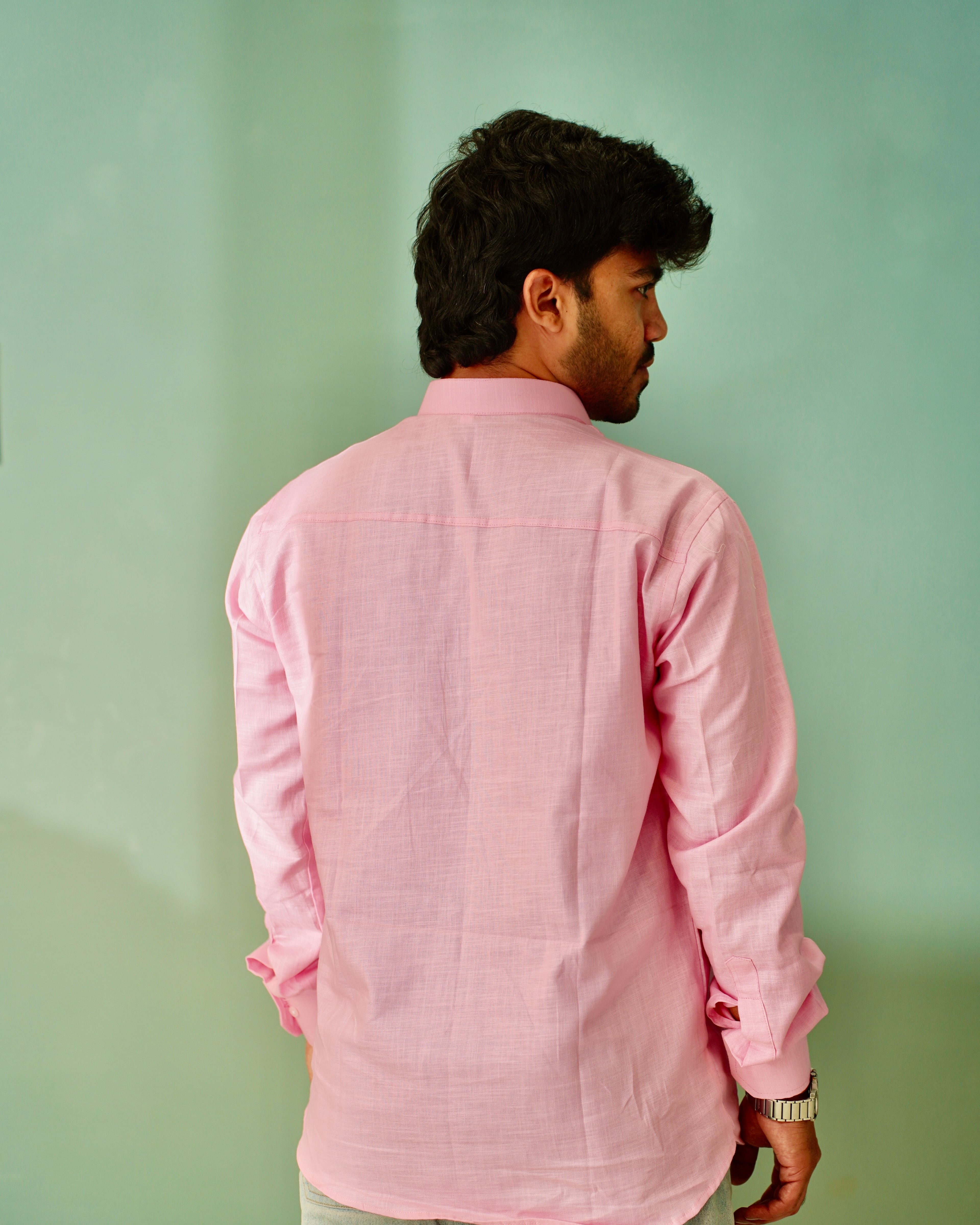 A man viewed from behind wearing a light pink long-sleeve linen shirt against a solid mint green background.