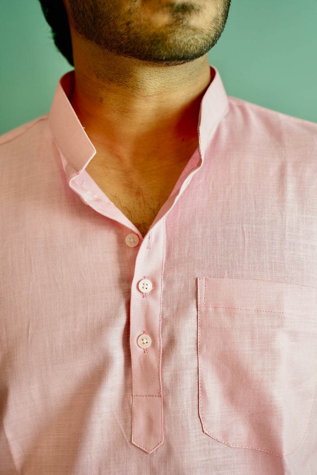 A close-up of a man wearing a light pink linen shirt with a spread collar, three-button placket, and a chest pocket, set against a solid mint green background.