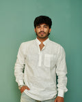 A man wearing a white long-sleeve linen shirt with a mandarin collar and chest pocket, standing against a solid sage green background.