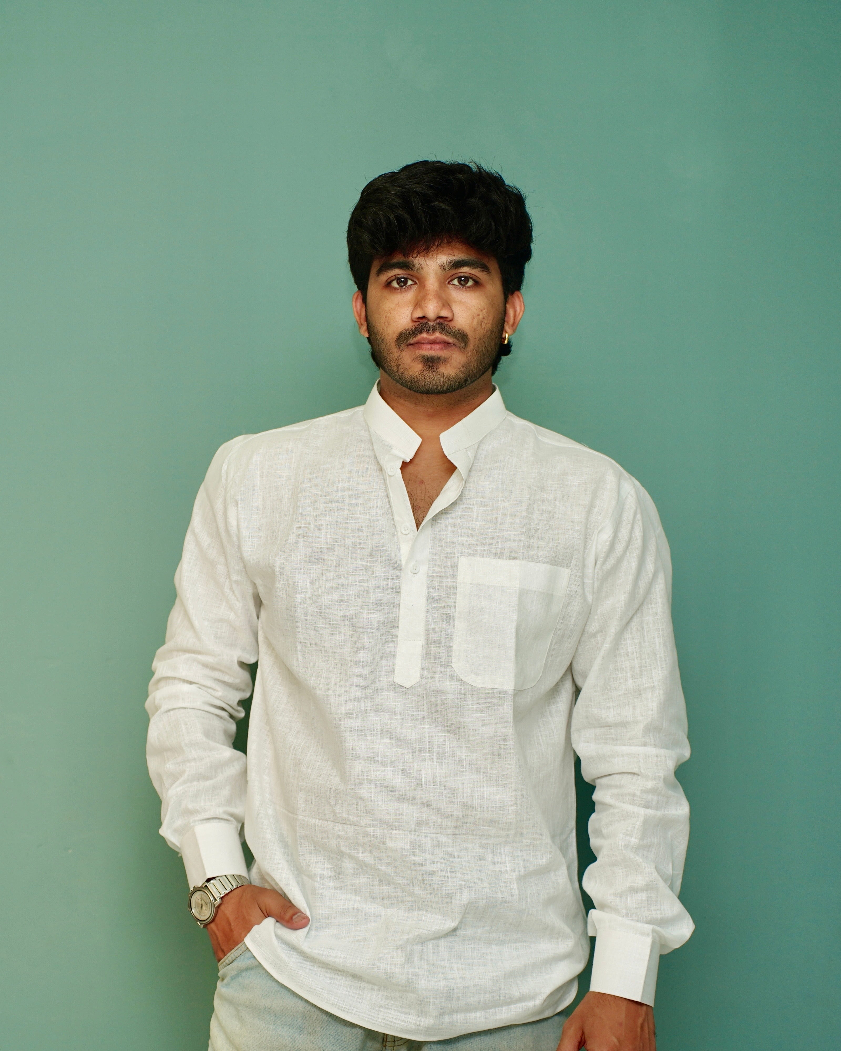 A man wearing a white long-sleeve linen shirt with a mandarin collar and chest pocket, standing against a solid sage green background.