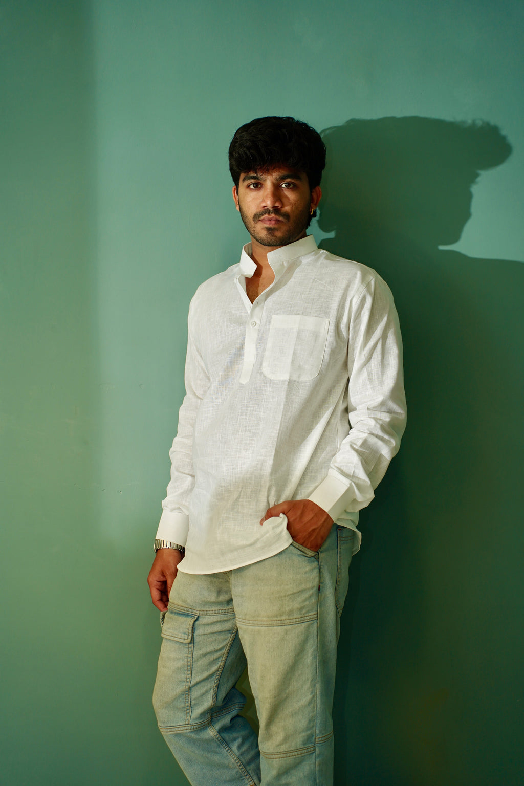 A man wearing a white long-sleeve linen shirt and light blue jeans, standing against a solid green wall with his hand in his pocket.