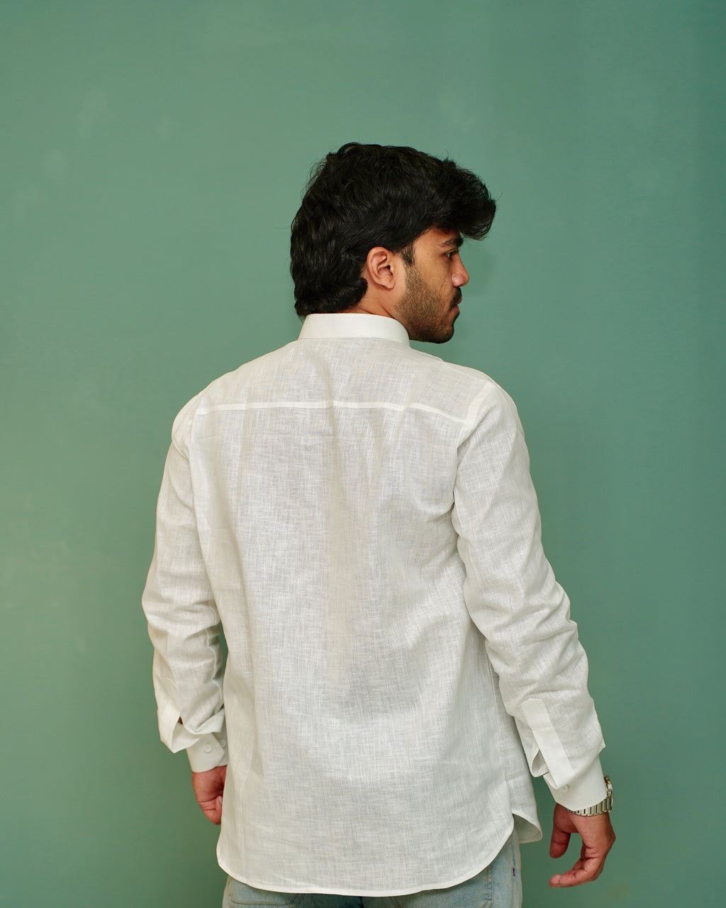 A man viewed from behind wearing a light beige linen shirt and light blue jeans, standing against a solid sage green background.