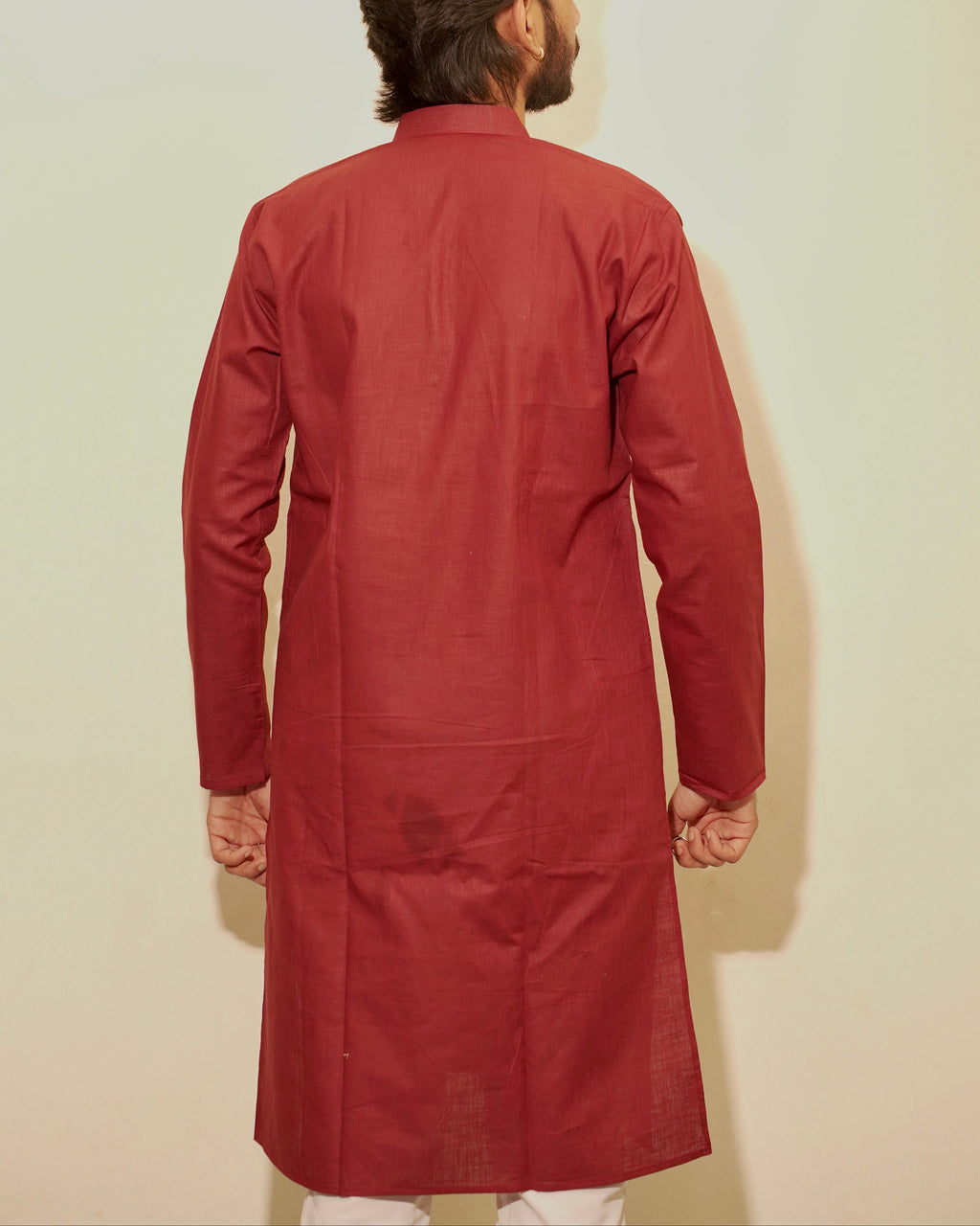 A man viewed from behind wearing a long-sleeved maroon kurta against a plain off-white background.