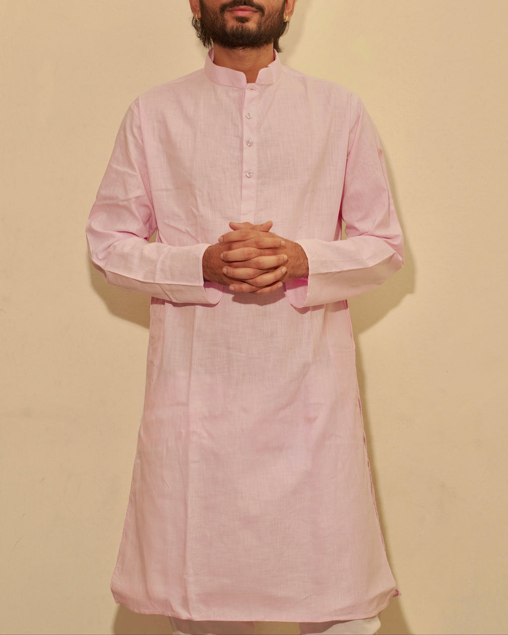 A man wearing a light pink long-sleeve kurta with a mandarin collar, standing against a plain beige wall with his hands clasped in front.