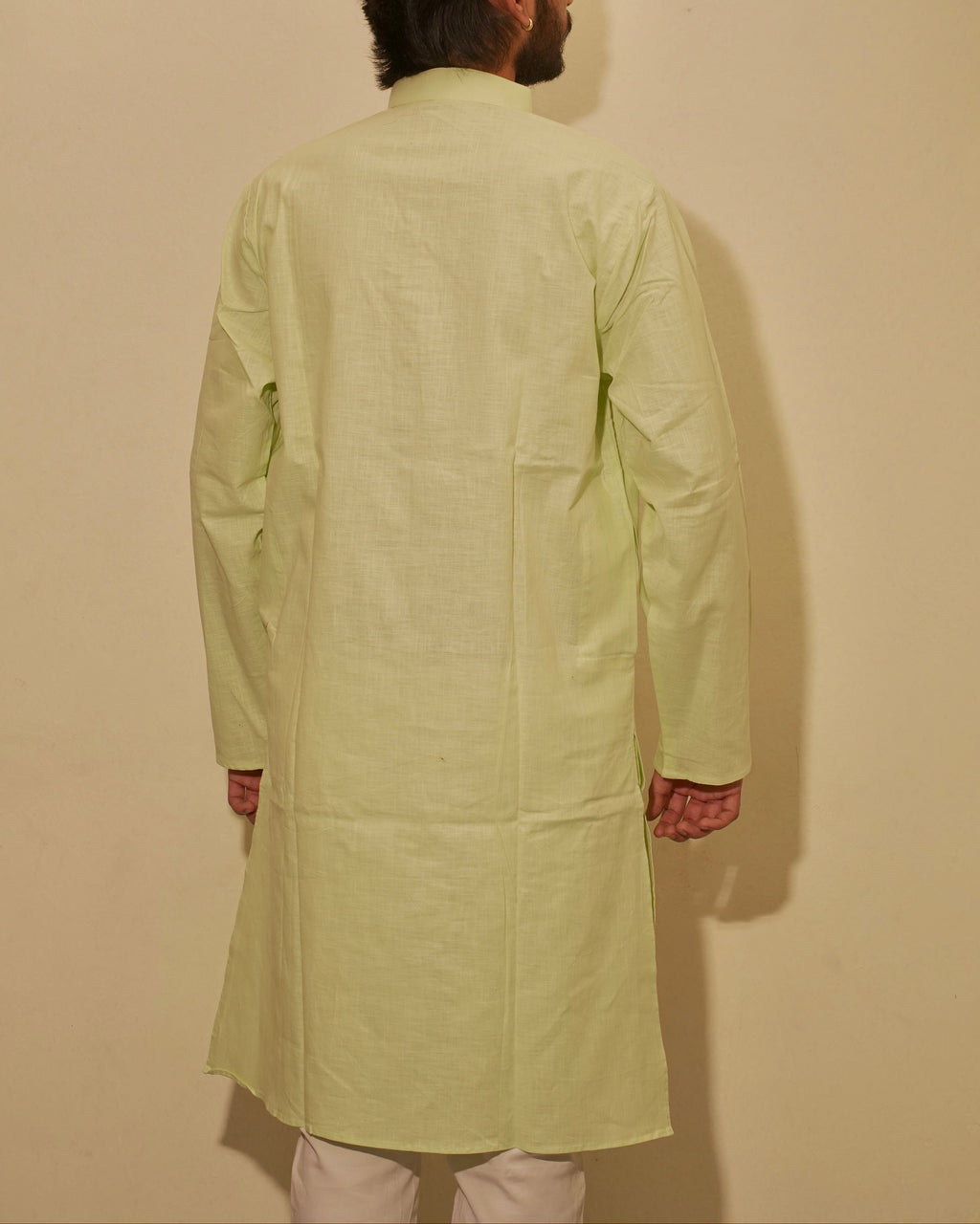 A man viewed from behind wearing a light sage green long-sleeved kurta and off-white trousers, standing against a plain beige wall.
