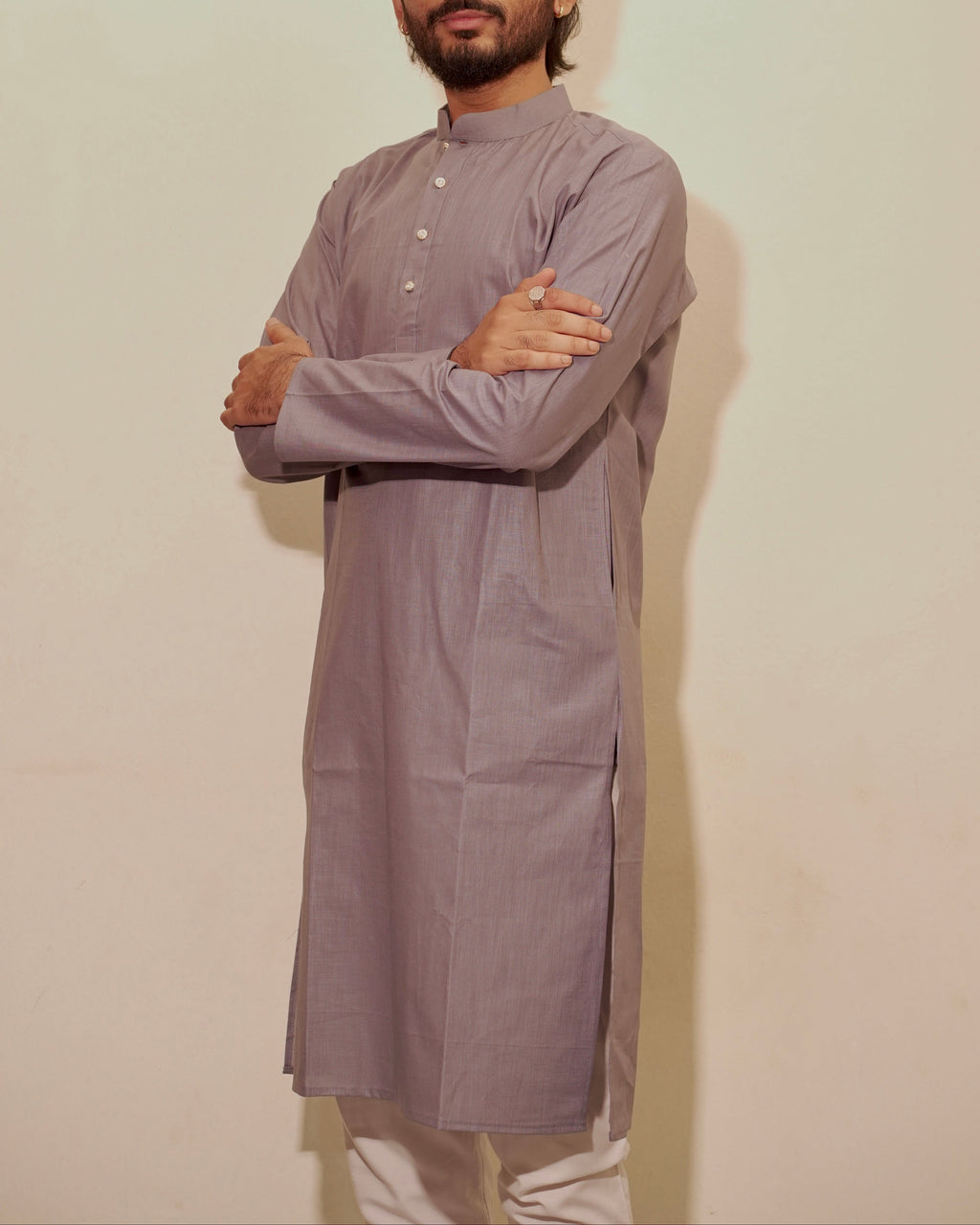A man with a beard modeling a gray long-sleeve kurta with white pants, standing against a plain off-white wall with his arms crossed.