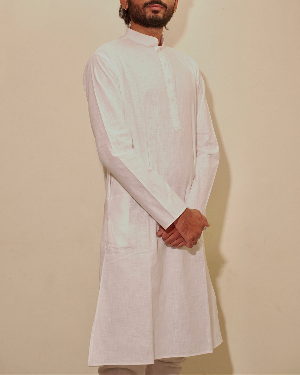 A man wearing a white long-sleeve kurta with a mandarin collar, standing against a plain beige wall.