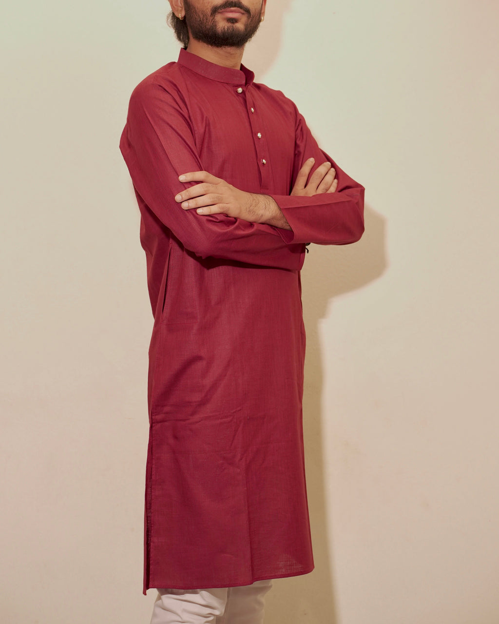 A man wearing a long-sleeved maroon kurta with white pajama, standing with arms crossed against a plain beige background.