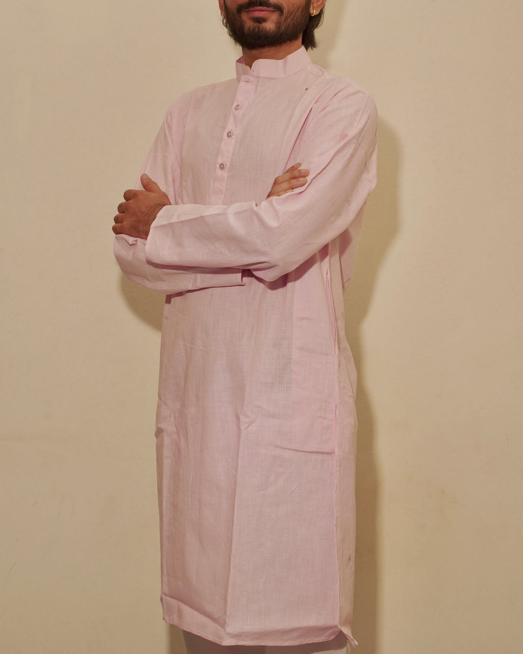 A man with a beard and short dark hair stands with arms crossed, wearing a light pink long-sleeved kurta against a plain beige wall.