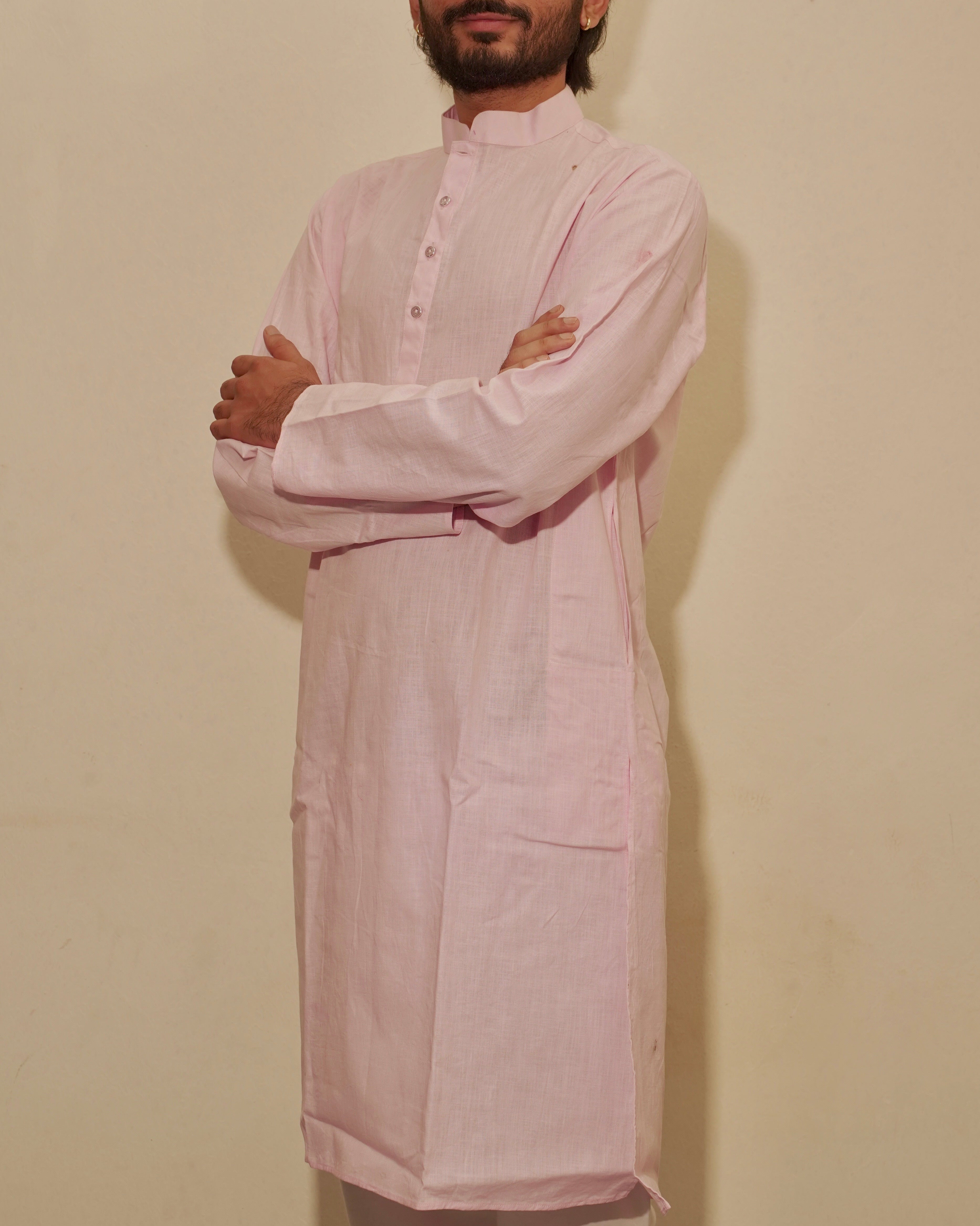 A man with a beard and short dark hair stands with arms crossed, wearing a light pink long-sleeved kurta against a plain beige wall.