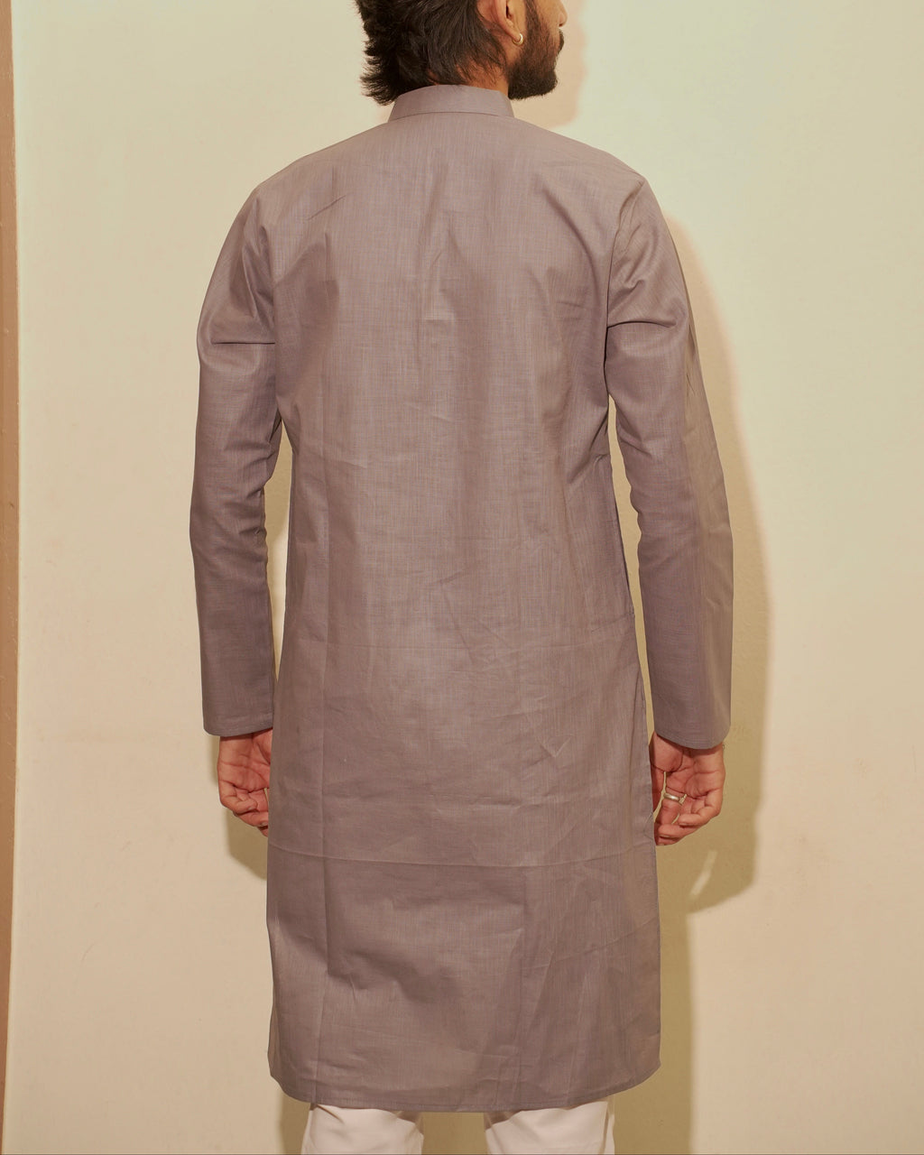 A man viewed from behind wearing a long-sleeved, knee-length light gray kurta against a plain beige wall.