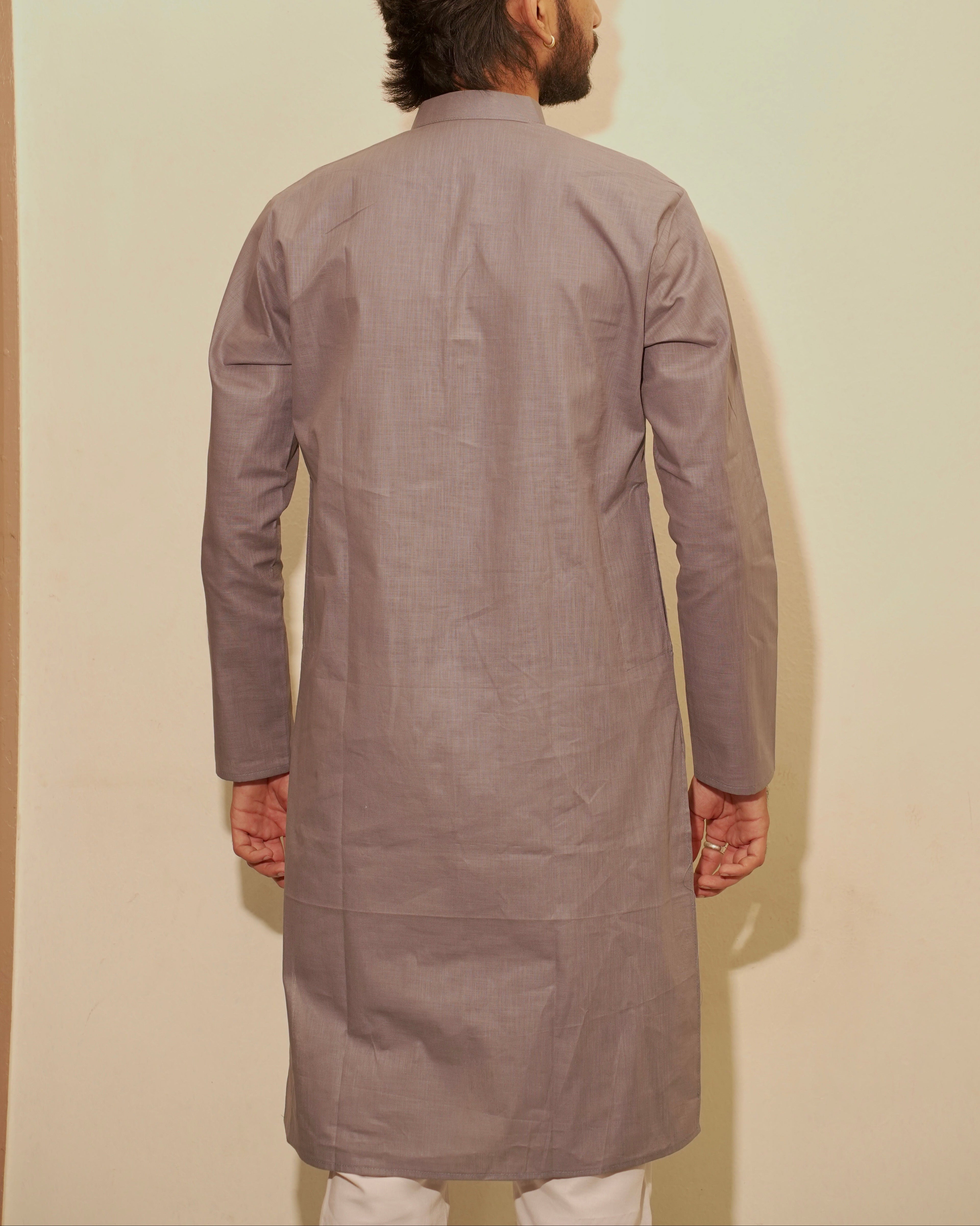 A man viewed from behind wearing a long-sleeved, knee-length light gray kurta against a plain beige wall.