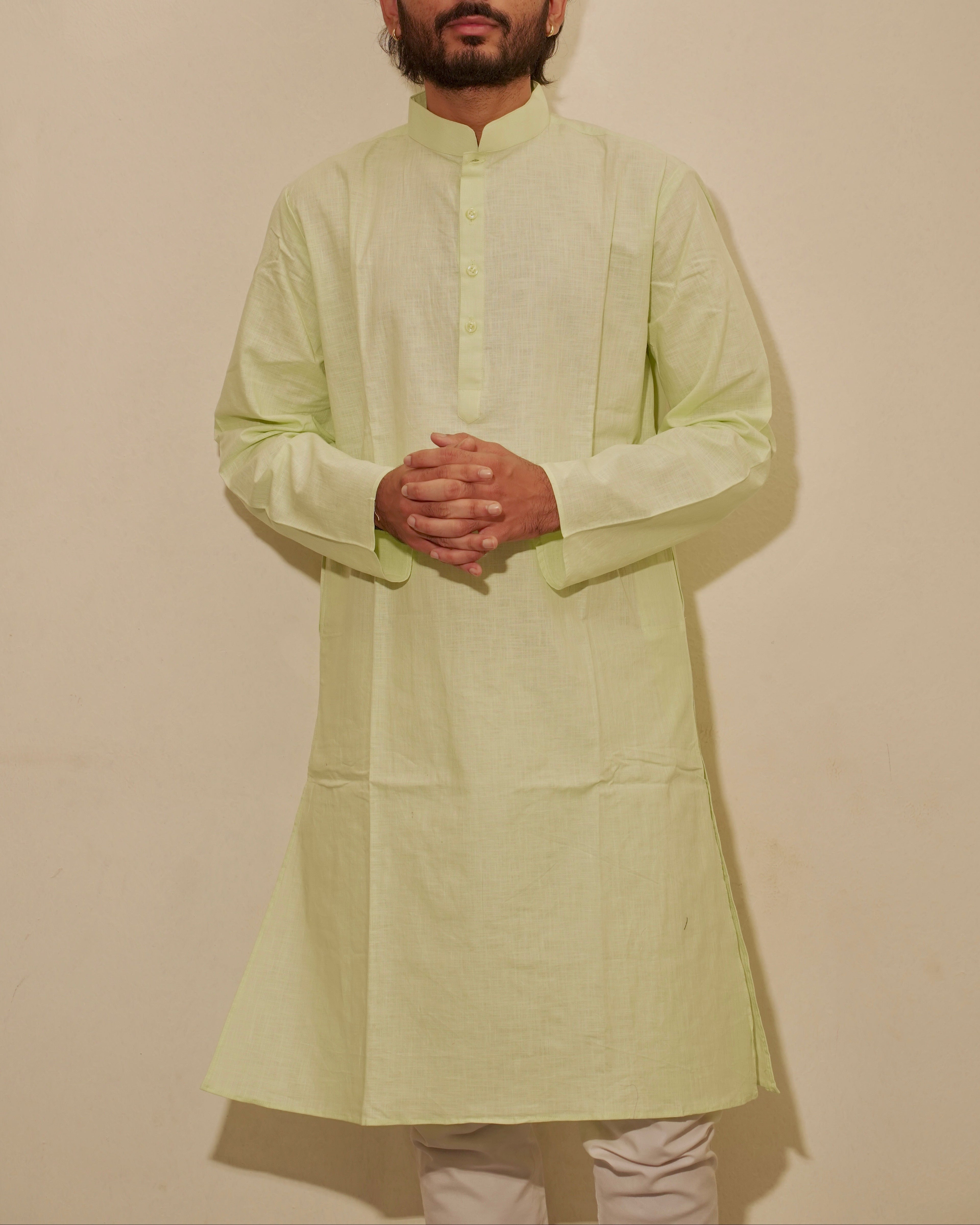 A man wearing a light lime green kurta with a mandarin collar and buttoned front, paired with light beige pajama, standing against a plain beige wall.