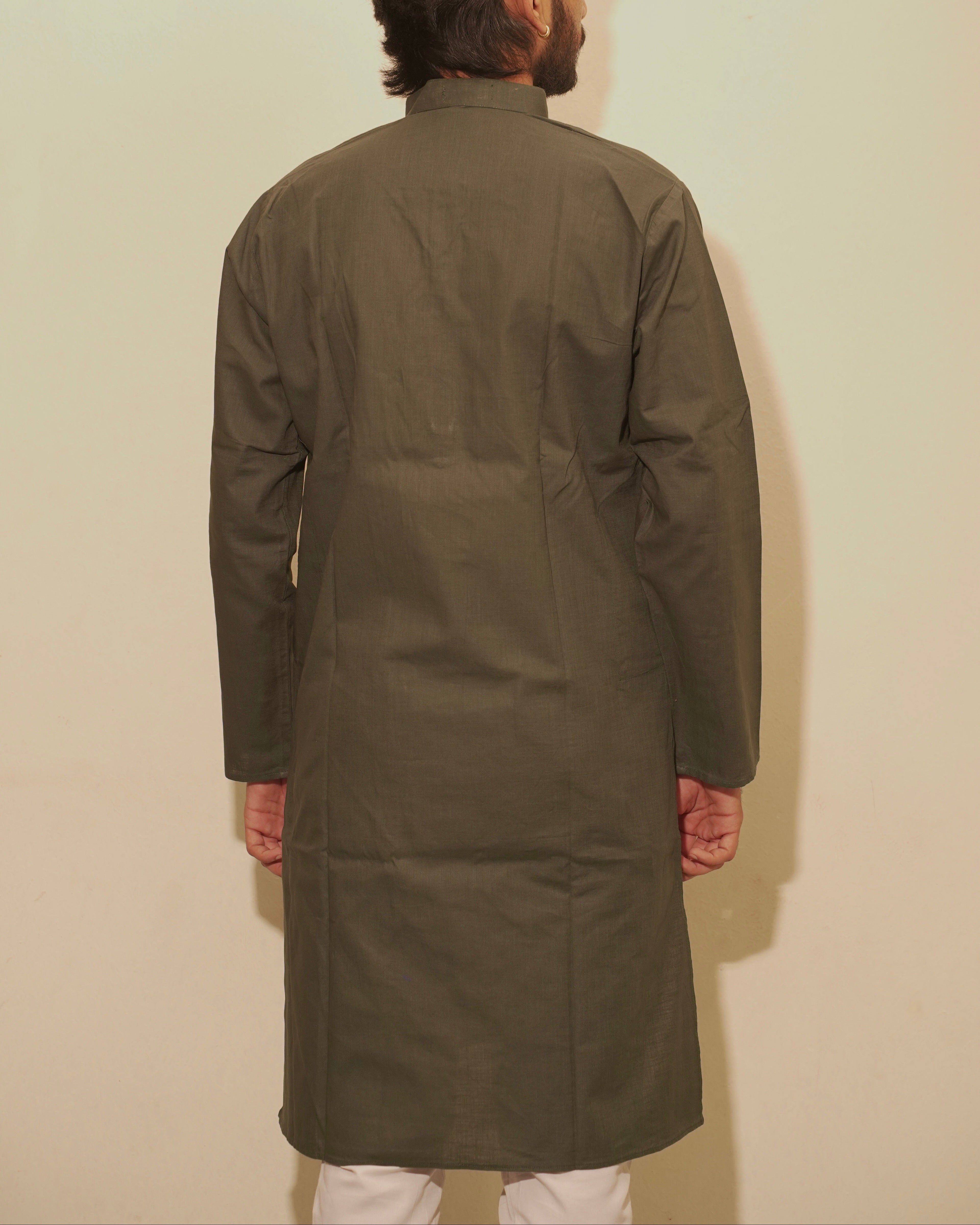 A man viewed from behind wearing a long-sleeved olive green kurta over a white shirt, standing against a plain light beige wall.