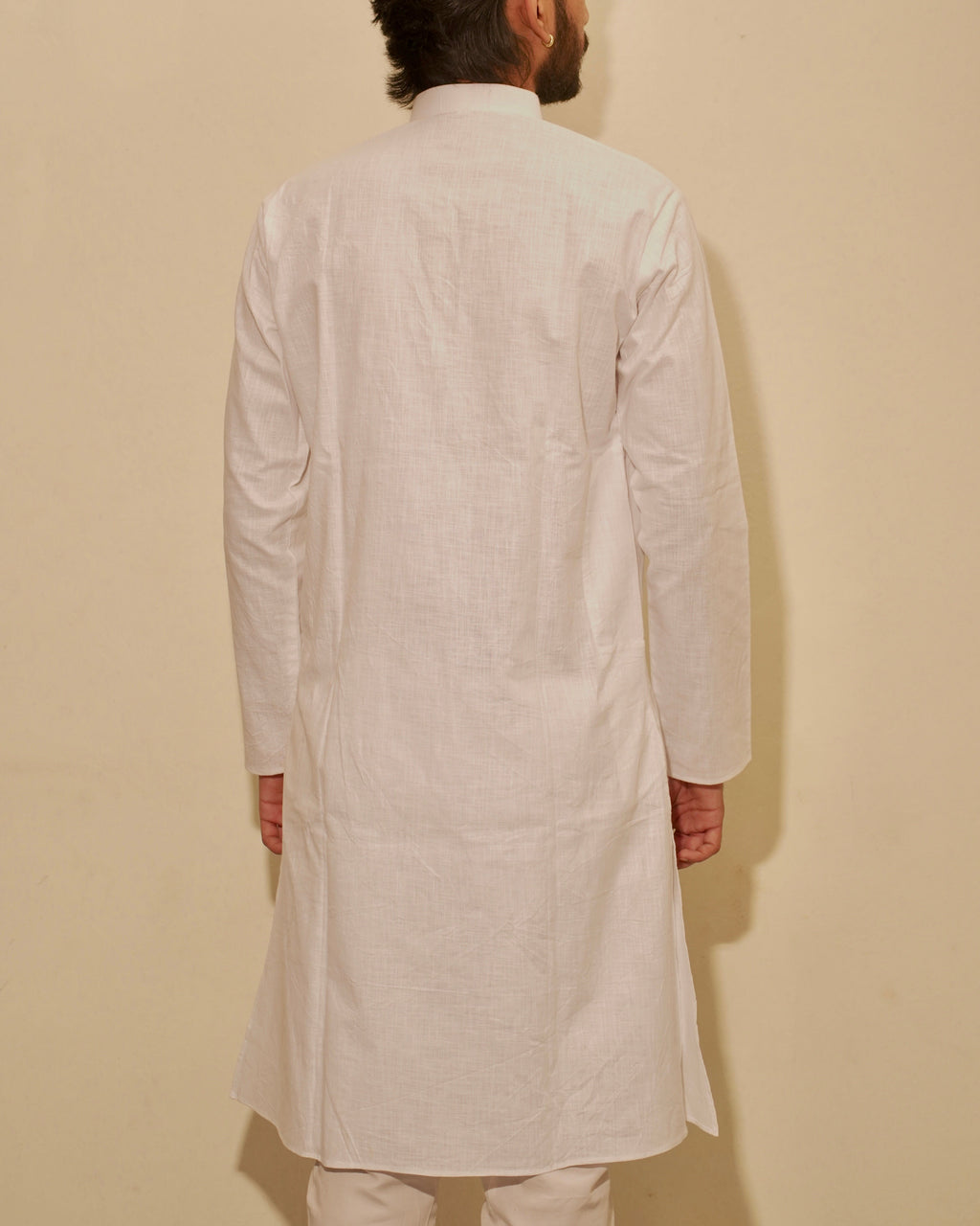 A man viewed from behind wearing a white long-sleeve kurta and matching pajama, standing against a plain beige wall.