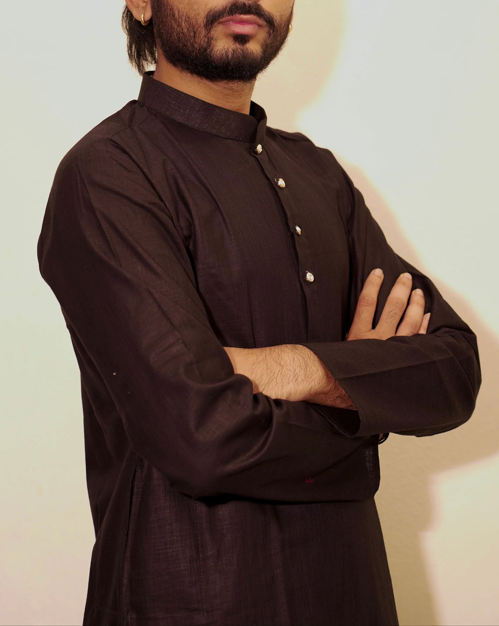 A man with a beard and styled hair wearing a Black long-sleeve button-up shirt, standing with arms crossed against a plain light background.