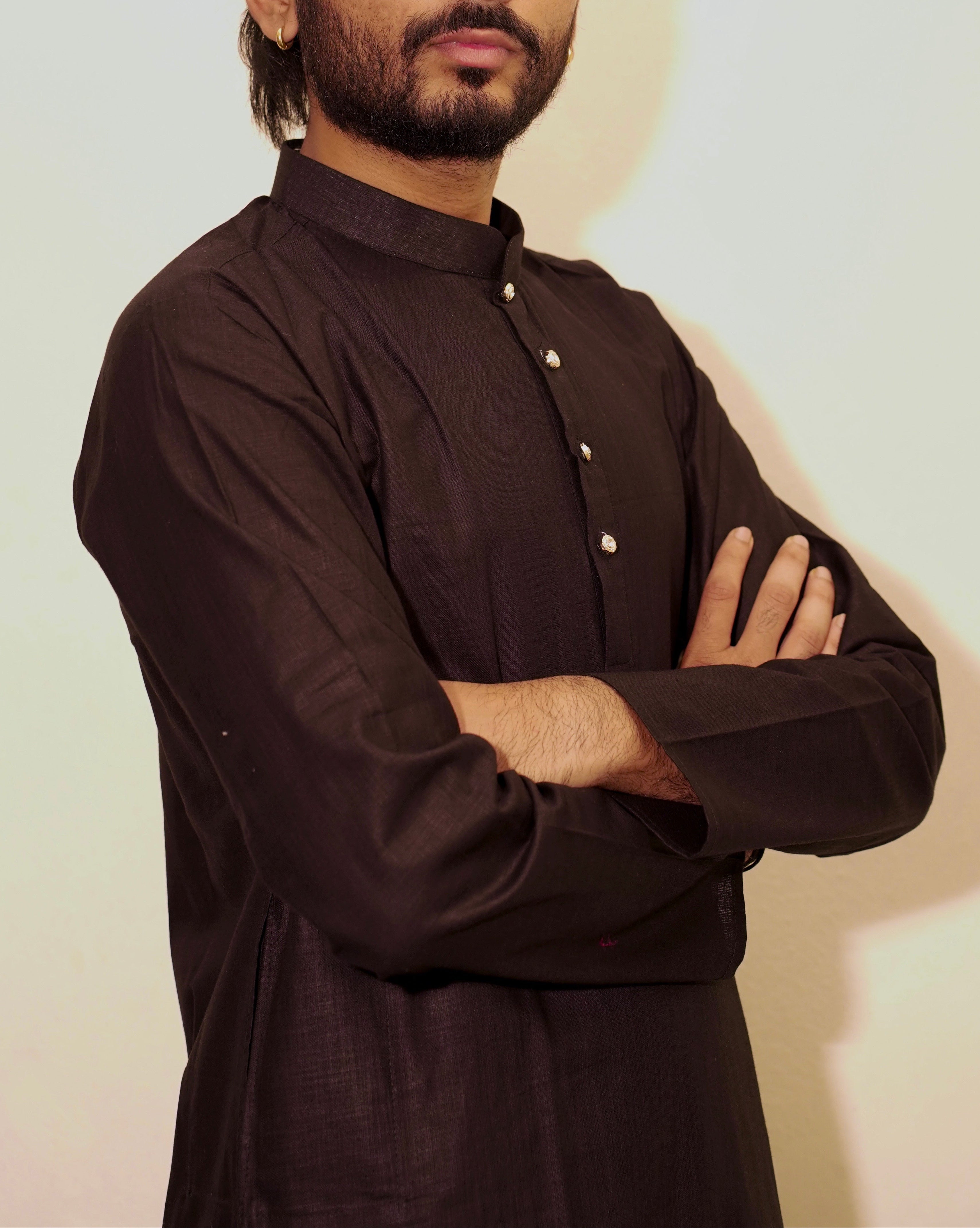 A man with a beard and styled hair wearing a Black long-sleeve button-up shirt, standing with arms crossed against a plain light background.