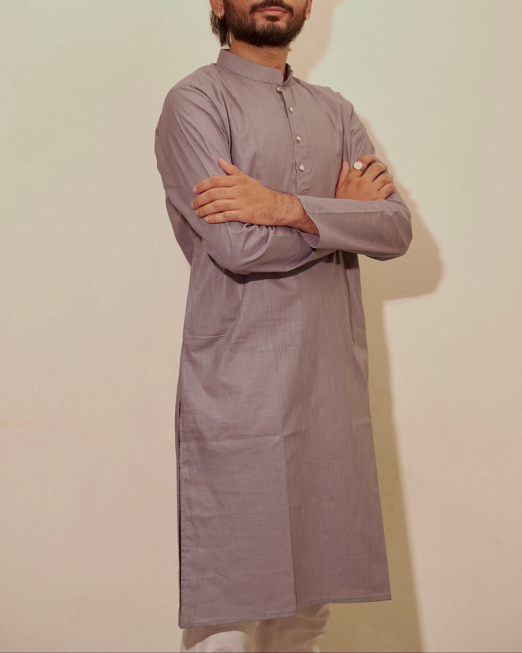 A man with a beard and styled hair stands wearing a long-sleeved, buttoned kurta in a muted gray color, posed with his arms crossed against a plain off-white background.