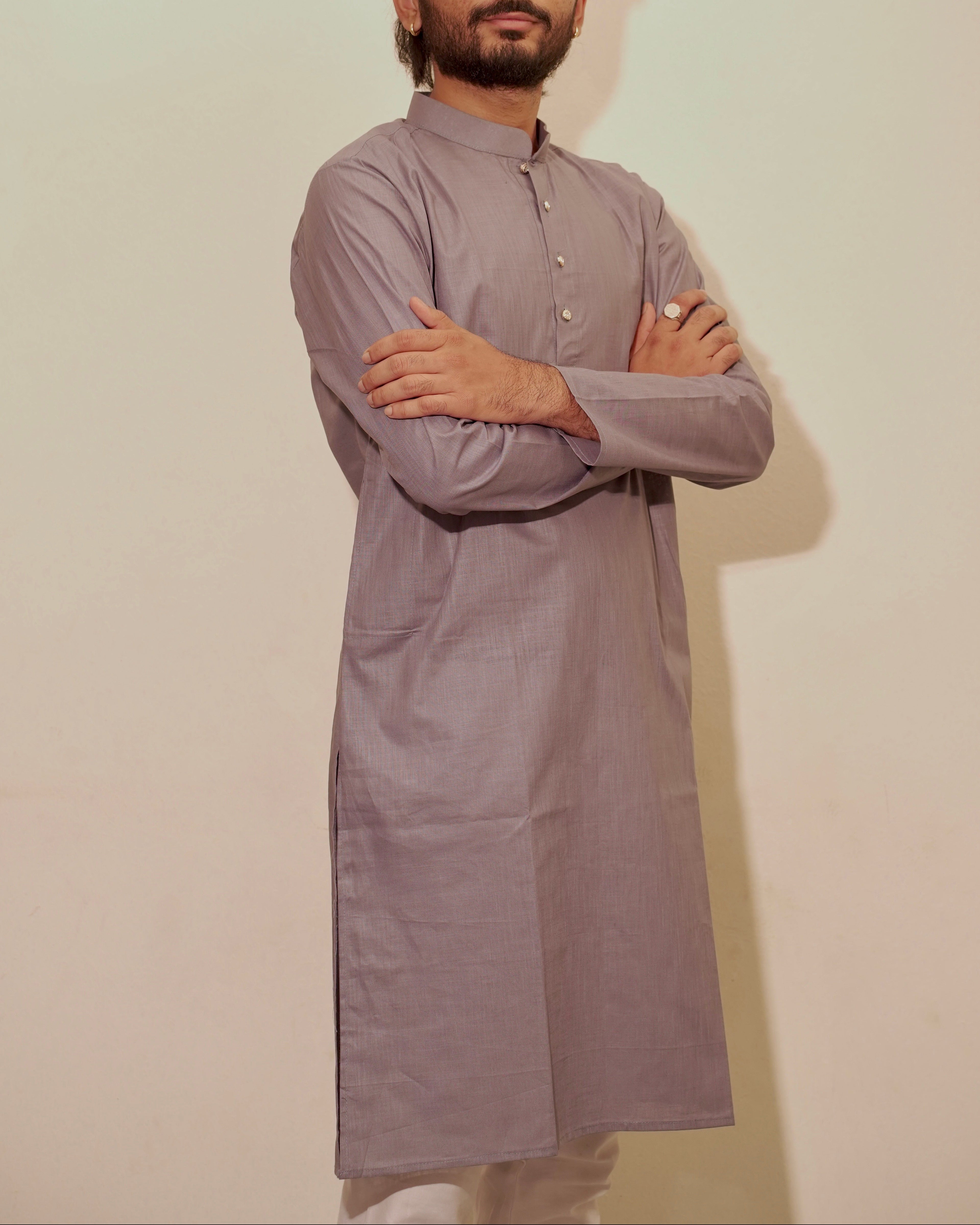 A man with a beard and styled hair stands wearing a long-sleeved, buttoned kurta in a muted gray color, posed with his arms crossed against a plain off-white background.