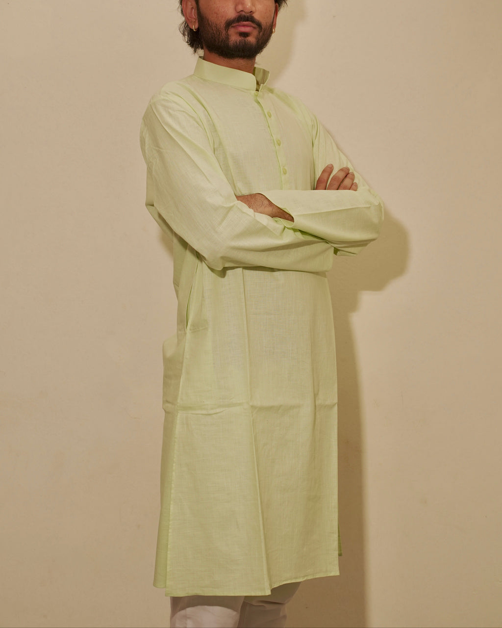 A man wearing a light lime green long-sleeve kurta with a mandarin collar, standing with arms crossed against a plain beige wall.