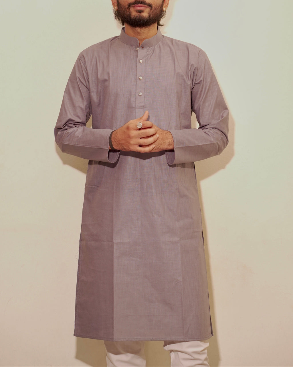 A man wearing a long-sleeved, knee-length gray kurta with a mandarin collar and button placket, paired with white pajama, standing against a plain off-white wall.