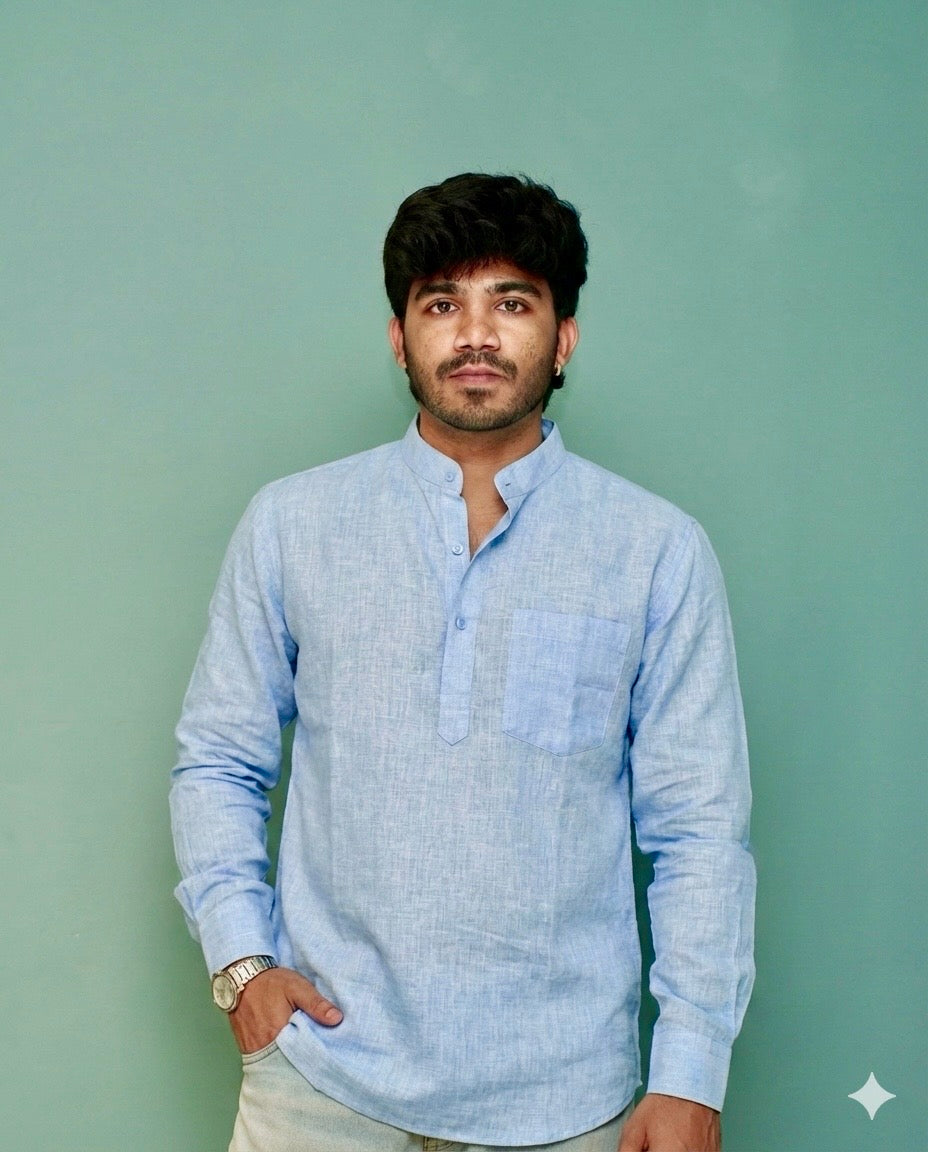 A man wearing a light blue long-sleeve linen shirt with a mandarin collar and a chest pocket, standing against a solid sage green background.